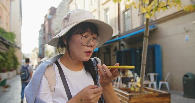 Close up portrait of happy middle-aged female tourist in hat and glasses standing on street and talking into speakerphone. Cheerful smiling Asian woman using virtual assistant app on cellphone outdoor