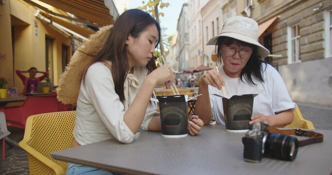 Portrait of joyful Asian mother and daughter sitting outdoor on restaurant terrace and eating chinese food. Young female having lunch with woman in cafeteria on street in city. Leisure concept