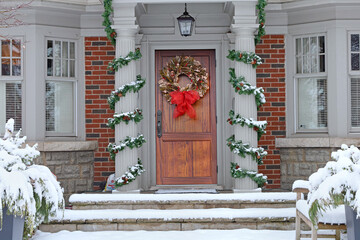 wood grain front door of house with colorful Christmas wreath with snow covered shrubs