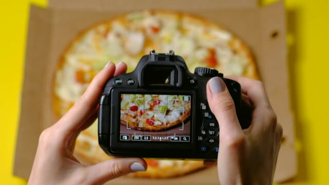  Top view - Female hands use dslr camera photographing appetizing fresh cooked italian pizza Caesar on yellow background. Food blogger of blogging concept.