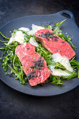 Modern style traditional fried New York strip steak with rucola and parmesan offered as close-up on a design plate