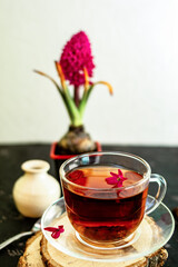 A cup of tea in a transparent cup with spring hyacinth flower on a black background. The mug stands on a wooden frame, next to it lies a metal spoon and a white vase. Copy space