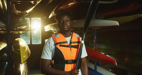 Portrait of African American serious male teen kayaker holding paddle in hand while standing indoors in warehouse with a stand with canoes. Young teenage boy looking at camera. Kayaking concept - Powered by Adobe