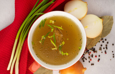 hot homemade beef bone broth with collagen and amino acids in red bowl with red napkin and green onions, black peas, chopped white onions and carrots on gray background. Concept support the immune