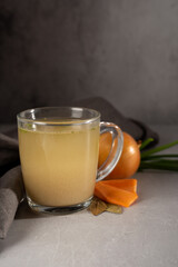 hot homemade chicken broth with collagen and amino acids in a transparent glass cup with gray napkin and green onions, black peas, white onions and carrots on gray background