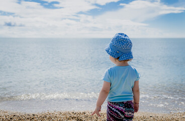 Fototapeta na wymiar Young girl standing on a beach and looking at a big blue sea. Baby girl at sea coast rearview.