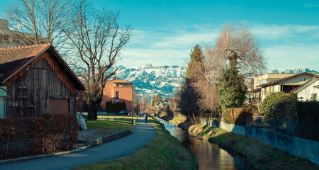 Fototapeta premium Alps mountains in Liechtenstein. Medieval Red House, calm narrow mountain river and jogging track, on the background of residential buildings, blue sky and snow-capped mountains. Liechtenstein, Vaduz
