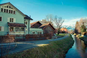Alps mountains in Liechtenstein. Medieval Red House, calm narrow mountain river and jogging track, on the background of residential buildings, blue sky and snow-capped mountains. Liechtenstein, Vaduz