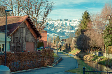 Alps mountains in Liechtenstein. Medieval Red House, calm narrow mountain river and jogging track, on the background of residential buildings, blue sky and snow-capped mountains. Liechtenstein, Vaduz