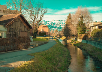 Fototapeta na wymiar Alps mountains in Liechtenstein. Medieval Red House, calm narrow mountain river and jogging track, on the background of residential buildings, blue sky and snow-capped mountains. Liechtenstein, Vaduz
