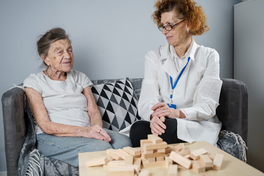 Dementia therapy in playful way, training fingers and fine motor skills, build wooden blocks into tower, playing Jenga. Senior woman 90 years old and doctor playing educational game in nursing home