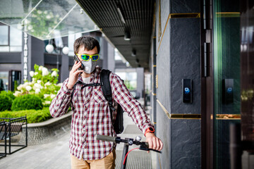 Fototapeta premium Courier young cute man using electric scooter delivered order to client on time, talking cell phone and waiting near front door of office, face mask protect by smog and car exhaust, delivery in city