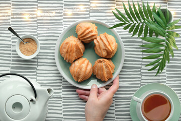 Creative flat lay with yummy eclairs or brewing cakes. White ceramic tea pot, cup of tee, sugar. Hand holds plate with cakes. Palm leaves in vase, light garland on light stripy textile tablecloth.