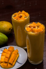 Mango lassi decorated with mango pulp raisin almonds and ripe mango in background mango slice in foreground