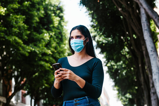 hispanic woman with facemask holding a phone and texting while she is walking in the street of a Latin America city