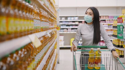 Young Asian woman in medical mask grocery walk toward camera shot, at supermarket during covid-19 coronavirus pandemic. Young lady stocks up food and toilet paper.
