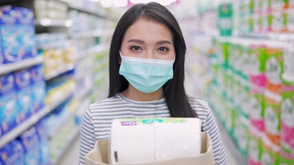 Young Asian woman in medical mask grocery walk toward camera shot, at supermarket during covid-19 coronavirus pandemic. Young lady stocks up food and toilet paper.