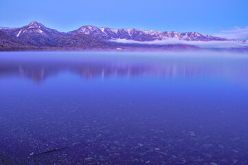 冬山と静水の湖面。夜明けの湖の風景。