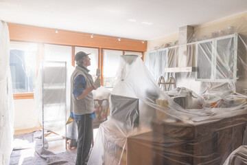 House painter painting the interior of a furnished house. Home renovation concept. Man is painting a kitchen protected with cover film