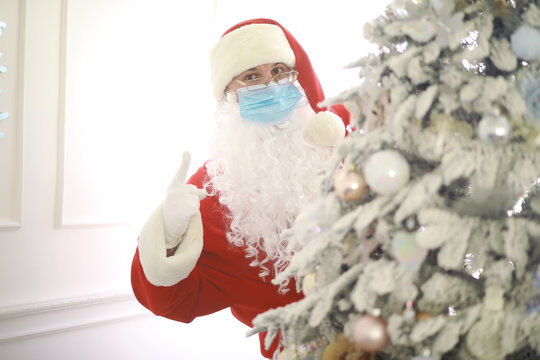 Real Santa Claus and christmas tree on a background, wearing a protective mask, glasses and hat. Christmas with social distance. Covid-19