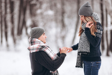 A guy kneeling down puts a wedding ring on a girl's hand, making a proposal to marry in a snowy...