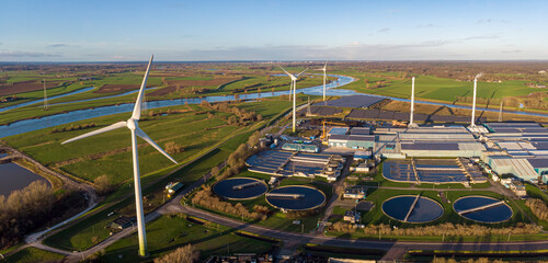 Wind turbines, water treatment and bio energy facility and solar panels in The Netherlands part of sustainable industry in Dutch flat river landscape against blue sky. Aerial circular economy concept.