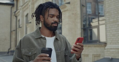 Close up view of cheerful guy with dreadlocks holding paper coffee cup and scrolling phone screen. Handsome bearded young man using smartphone while walking at street - Powered by Adobe