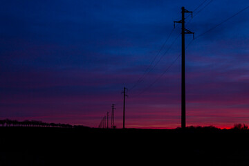 Fototapeta na wymiar A silhouette of a line of electric poles with cables of electricity against the backdrop of a colorful sunset pink purple blue cloudy sky.