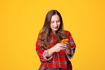 Happy young female in checkered shirt smiling and browsing social media on smartphone against yellow background