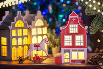 festive composition of lamps in shape of old European houses, toy horse and pine cones, against a background of garlands in defocus.