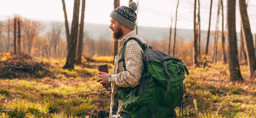 Bearded man with backpack and drink to go travelling through forest in autumn in nature
