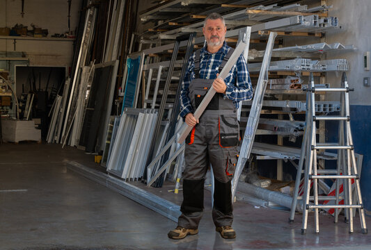 Portrait of an elderly mechanic. The experienced man with a beard is wearing work trousers and has an aluminum profile in his hands. In the background are shelves for windows and terrace construction. - Powered by Adobe