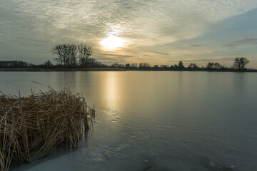 Frozen lake with dry reeds, sunset and clouds on the sky