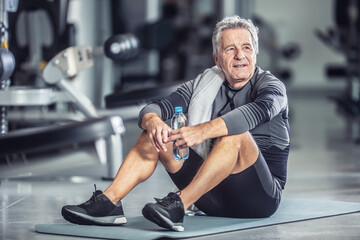 Fototapeta na wymiar Older male rests with a bottle of water sitting on a mat during a gym workout