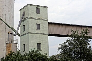 part of an old cement factory made of a brick tower with windows and a gray wooden structure against a sky overgrown with green vegetation