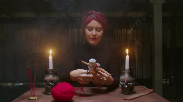 Senior witch woman fortune teller uses voodoo doll and red rope to terrible magic ritual