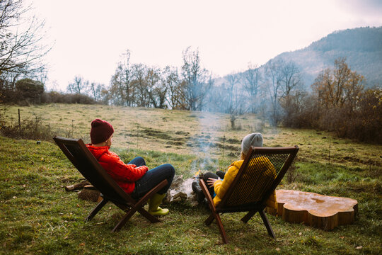 Couple in warm puffy jackets sit on sun chairs in front of cozy campfire at campground or country house. Off grid living in nature. Millennial weekend getaway to camp overnight location