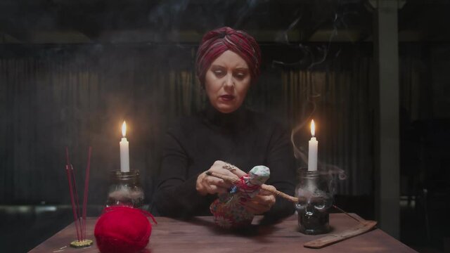 Senior witch woman fortune teller sticks a needle into voodoo doll in terrible magic ritual