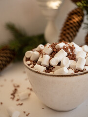A cup of cocoa with marshmallows on the background of fir branches with cones in a vase and a burning candle on a light background in the morning light