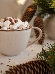 A cup of cocoa with marshmallows on the background of fir branches with cones in a vase and a burning candle on a light background in the morning light