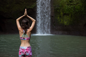 Hands raising up in namaste mudra. Woman meditating, practicing yoga and pranayama with namaste mudra near waterfall. Yoga outdoor concept. Tibumana waterfall, Bali. Copy space. View from back.