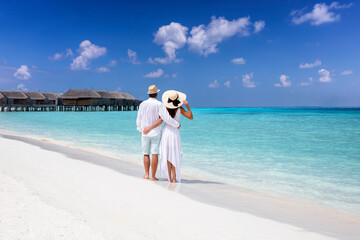 A hugging couple in white summer clothing stands on a tropical beach and enjoys the view to the...