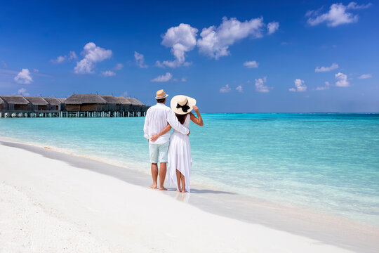 A hugging couple in white summer clothing stands on a tropical beach and enjoys the view to the turquoise sea and blue sky - Powered by Adobe