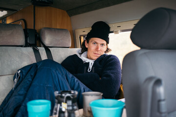 Handsome young man, traveller on road trip, sit inside camping van on rainy day. Cosy comfortable setup in custom camper trailer or van. Millennial travel trend, adventure on the road. Cosy camping