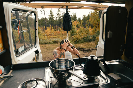View from inside camper van on young hipster man take shower outdoors from camping pocket shower. Life inside converted camping van on the road. Vanlife lifestyle concept. Funny camping