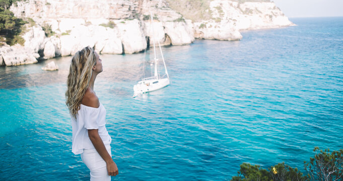 Carefree female tourist breathing freedom during summer vacations in Spanish resort with Mediterranean coastline, youthful traveler in white clothing enjoying picturesque views of Menorca island