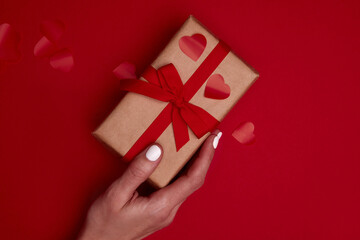 Valentine day composition with copy space. Woman hand holding present or gift box wraped in craft paper with ribbon bow and red hearts confetti. Close up, top view, copy space