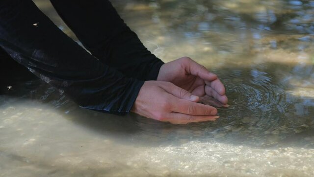 Close up detail thirsty wanderer greedily drinking life-giving water from a stream or lake. Hands of long-haired man in dirty dark clothes  collects clear water from the lake. 200 fps slow motion.