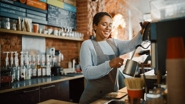 Beautiful Latin American Female Barista with Short Hair and Glasses is Making a Cup of Tasty Cappuccino in Coffee Shop Bar. Male Cashier Works at a Cozy Loft-Style Cafe Counter in the Background.