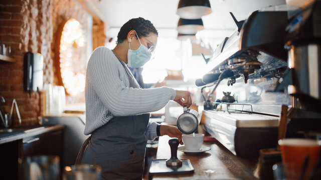 Beautiful Latin American Female Barista in Face Mask is Making a Cup of Cappuccino in Coffee Shop Bar. Social and Medical Health Restrictions Concept in a Loft-Style Cafe During Coronavirus Pandemic.